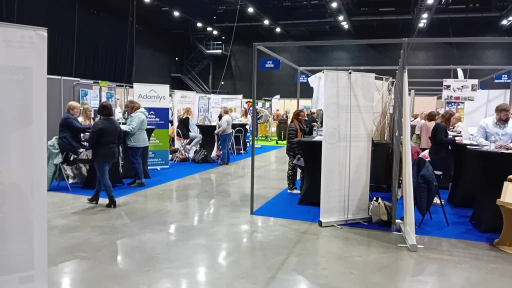 Vue intérieure du Salon du Travail de Saint-Omer 2025 avec plusieurs stands d’entreprises et d’organismes, installés sur moquette bleue.